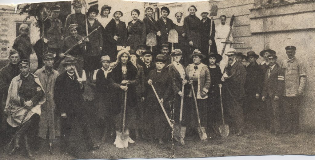 Czarno-białe zdjęcie grupowe kobiet i mężczyzn stojących w dwóch rzędach. Postaci stojące w drugim rzędzie widziane są do kolan. Pozują cywile i kilkoro żołnierzy w płaszczach. W pierwszym rzędzie pięć kobiet w sukniach, lekkich okryciach wierzchnich i kapeluszach pozuje z łopatami w rękach.