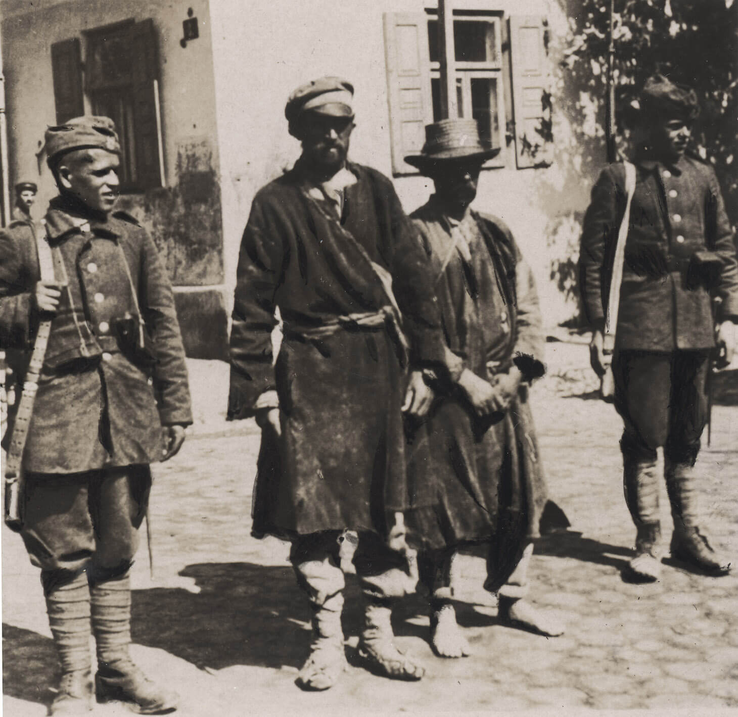 Czarno-białe zdjęcie plenerowe przedstawiające dwóch bolszewików pilnowanych przez dwóch polskich żołnierzy. Na pierwszym planie stoją od lewej: uśmiechnięty żołnierz polski w mundurze polowym, z karabinem przewieszonym przez ramię. Drugi od lewej, stoi jeniec z opuszczonymi rękami. Ubrany jest w ciemny, obdarty, brudny, płaszcz chłopski, przepasany powrozem. Na nogach ma bezkształtne owijacze, na głowie czapkę z daszkiem. Mężczyzna patrzy w obiektyw. Trzeci od lewej w podobnym ubraniu, stoi boso ze złączonymi z przodu rękami, w słomkowym kapeluszu na głowie. Czwarty od lewej, to drugi polski żołnierz, także w mundurze polowym, z karabinem z bagnetem przewieszonym przez ramię. Pozujący stoją na bruku z kamieni. W tle widać narożnik domu wiejskiego z oknami z otwartymi okiennicami.

