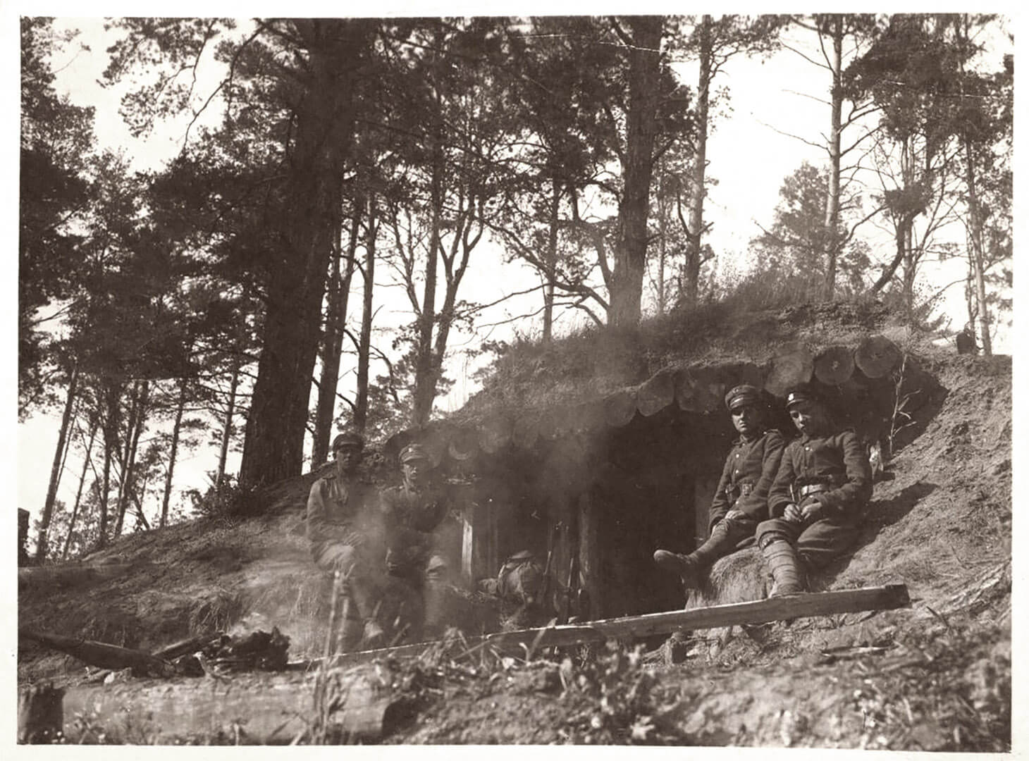 Czarno-białe zdjęcie plenerowe pokazujące czterech żołnierzy pozujących przed umocnieniem w lesie. Zdjęcie wykonane z niskiej perspektywy ukazuje centralnie schron o konstrukcji ziemno-drewnianej. Po lewej i prawej stronie otworu-wejścia siedzi swobodnie na piaszczystym nasypie po dwóch polskich żołnierzy. Mężczyźni patrzą w obiektyw. Za umocnieniem widać iglaste zalesienie.