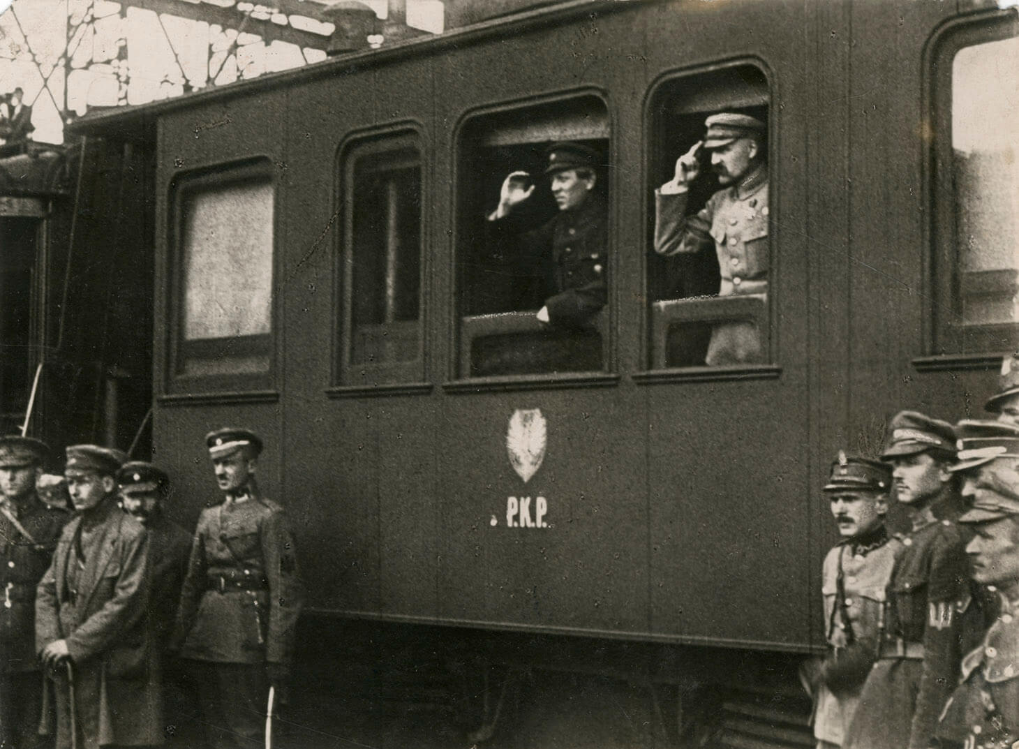 Czarno-biała fotografia przedstawia Naczelnego Wodza Józefa Piłsudskiego i atamana Symona Petlurę, stojących w oknach wagonu kolejowego i salutujących. Okna, które znajdują się w centrum kadru, są opuszczone. W oknie pierwszym od lewej ataman Petlura ubrany w ciemny mundur, w drugim oknie  Naczelny Wódz Józef Piłsudski w jasnym mundurze i czapce maciejówce. Pod oknami, na wagonie namalowane jest godło z orłem i napis P.K.P. Przed wagonem, na peronie stoją swobodnie oficerowie kierując wzrok w tę samą stronę co główni bohaterowie zdjęcia.

