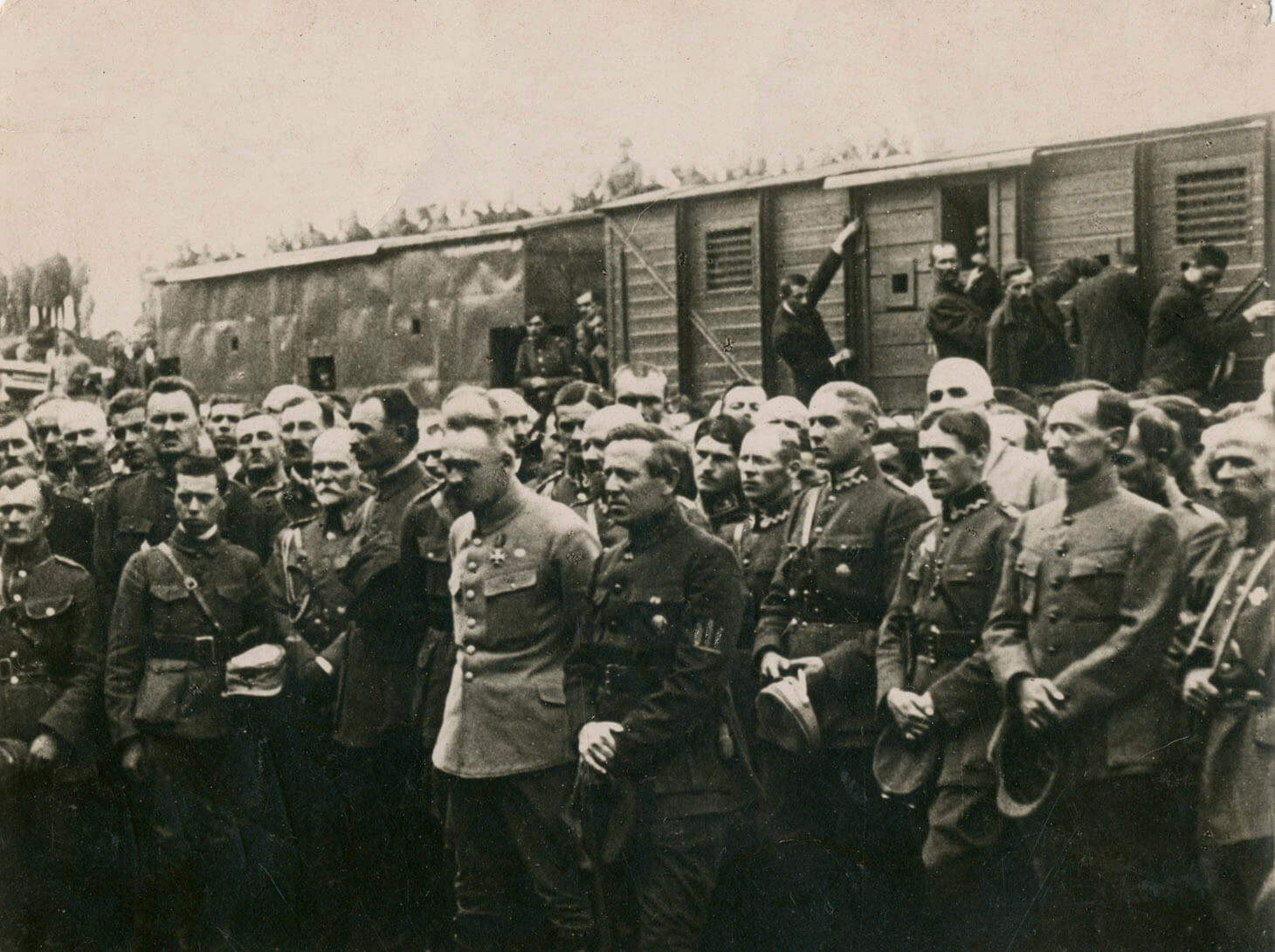 Czarno-biała fotografia grupowa tłumu żołnierzy na tle składu kolejowego. Na pierwszym planie, w centrum kadru znajdują się Naczelny Wódz Józef Piłsudski oraz . Ubrany jest w jasną kurtkę mundurową i ciemne spodnie, bez nakrycia głowy.o oraz drugi od lewej: ataman Symon Petlura w ciemnym mundurze. Za nimi stoją oficerowie. Wszyscy zebrani pokazani są  bez nakryć głowy, które trzymają w swobodnie opuszczonych, złączonych przed sobą rękach. 

