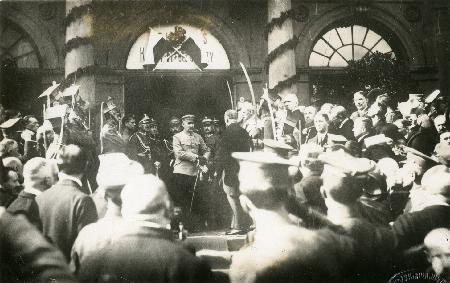 Czarno-biała fotografia ukazuje Marszałka Józefa Piłsudskiego na tle szerokich drzwi dworca, nad którymi przeszklone półkole prześwitu udekorowane jest skrzyżowanymi, polskimi flagami. Dwie kolumny zdobiące wejście są owinięte girlandami. Marszałek w jasnej kurtce mundurowej i ciemnych spodniach, w czapce  maciejówce, z szablą przy pasie, wychodzi w otoczeniu oficerów. Przed nimi, po obydwu stronach utworzony jest szpaler kawalerzystów w wysokich rogatywkach ułańskich, z szablami w chwycie “Prezentuj”. Na pierwszym planie, nieostre sylwetki żołnierzy i cywilów stojących tyłem do widza.
