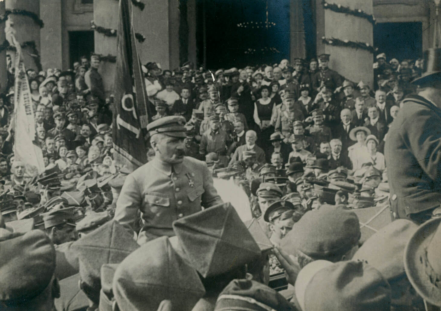 Czarno-biała fotografia ukazująca Naczelnego Wodza Józefa Piłsudskiego przed wejściem do kościoła św. Aleksandra na Placu Trzech Krzyży otoczonego przez tłum żołnierzy i cywilów. W tle widoczne jest wejście do świątyni, a także udekorowane kolumny po obydwu stronach wejścia. Nad tłumem unoszą się dwie chorągwie z nieczytelnymi emblematami. Centralnie, ponad głowami innych, wyróżnia się widziana do pasa, postać Marszałka Józefa Piłsudskiego. Wódz pokazany jest w jasnym mundurze i czapce maciejówce, z orderem po lewej stronie piersi. Tłum otacza bryczkę, w której Marszałek siedzi. Po prawej stronie widoczny jest stangret, kierujący pojazd w prawą stronę kadru. Na pierwszym planie znajdują się głowy w czapkach rogatywkach i kapeluszach, widziane od tyłu.