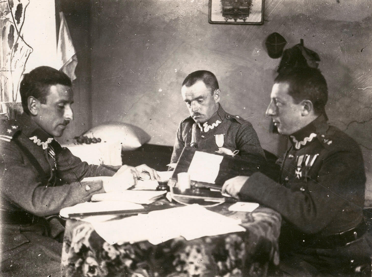 Zdjęcie we wnętrzu mieszkalnym. Trzech oficerów siedzi przy okrągłym stole. Światło wpada przez okno po lewej stronie kadru. 
Na pierwszym planie widać blat z kwiecistym obrusem, na nim rozłożone papiery. Pierwszy od lewej, siedzi prawym bokiem do obiektywu,  adiutant pułkownika Stanisława Skwaczyńskiego. Żołnierz ubrany jest w mundurze spięty pasem z koalicyjką. Ręce ma oparte o krawędź stołu, w prawej dłoni trzyma pióro. Drugi od lewej siedzi pułkownik Stanisław Skwarczyński. W ujęciu, w lekkim zwrocie w prawo, czyta jakiś dokument. Trzeci od lewej, pokazany jest drugi adiutant pułkownika. Żołnierz pokazany jest z lewego boku. Trzyma lewą rękę opartą o krawędź stołu i patrzy na wprost, na swojego kolegę. W głębi kadru, po lewej stronie, w narożniku pomieszczenia, widać poduszkę, leżącą na małej kanapie. Nad nią, na ścianie wisi obraz lub lustro w ramce. Po prawej stronie, na ścianie wisi czapka rogatywka i płaszcz wojskowy.
