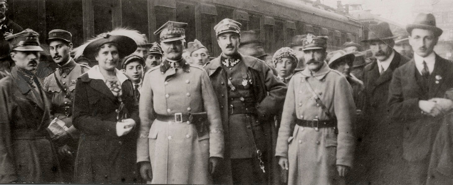 Czarno-białe, grupowe zdjęcie panoramiczne, wykonane na peronie kolejowym. W tle widać wagon pociągu pasażerskiego. Do zdjęcia pozują, stojąc na wprost obiektywu, w ujęciu do kolan: pierwszy od lewej, generał Mieczysław Kuliński w płaszczu wojskowym i rogatywce, drugi, pułkownik Stanisław Dowoyno-Sołłohub, w rozpiętym płaszczu wojskowym i rogatywce, trzeci kapitan Jaworowski, także w płaszczu wojskowym i czwarty, doktor Frenkner, w płaszczu i kapeluszu. Mężczyźni stoją w otoczeniu innych żołnierzy, kobiet i cywilów.