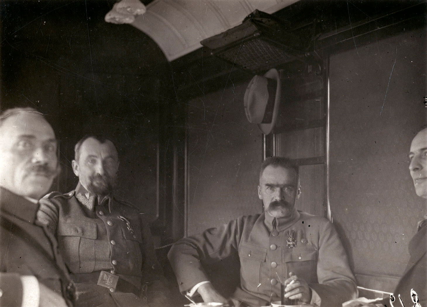 Czarno-białe zdjęcie Marszałka Piłsudskiego i Generała Tadeusza Rozwadowskiego w towarzystwie dwóch mężczyzn w wagonie pociągu. Mężczyźni pokazani są w planie półpełnym czyli do pasa. Wnętrze wagonu obite jest dekoracyjną tkaniną. Pierwszy od lewej, na mężczyzna na pierwszym planie, z zarostem pod nosem, siedzi prawym bokiem do obiektywu, w którego stronę spogląda. Drugi od lewej, w drugim planie, siedzi pokazany en face Generał Tadeusz Rozwadowski. Trzeci od lewej, pokazany jest en face Marszałek Józef Piłsudski. Wódz podpiera się prawą ręką pod bok, lewą z papierosem w dłoni, opiera o stolik. Czwarty mężczyzna, widoczny jest częściowo, lewym półprofilem. Jego postać przycięta jest prawym spadem zdjęcia. Za Marszałkiem Piłsudskim, na ścianie  wagonu znajduje się stylowy wieszak, na którym zawisa jasny kapelusz w stylu Homburg.