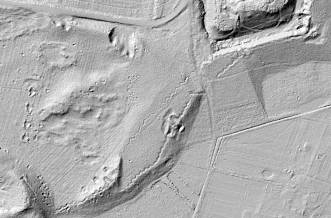 Czarno-białe zdjęcie ukazujące zarys umocnień Fortu Beniamin w ujęciu z lotu ptaka. Abstrakcyjny obraz przedstawia jakby odciśnięte w piasku linie, wężyki i wgniecenia, które odpowiadają miejscu umocnień. Wężyki biegną po przekątnej od lewego, dolnego rogu. W prawym, dolnym rogu linie proste otaczają róg kadru pod kątem mniej więcej 120 stopni.

