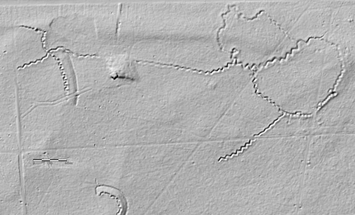 Czarno-białe zdjęcie ukazujące zarys północno-wschodniego przedpola Twierdzy Modlin z lotu ptaka. Abstrakcyjny obraz przedstawia jakby odciśnięte w piasku linie, wężyki i wgniecenia, które odpowiadają miejscu umocnień. Uwagę zwraca, usytuowany u góry kompozycji, szlak utworzony z wężykowatej, zygzakowatej linii.

