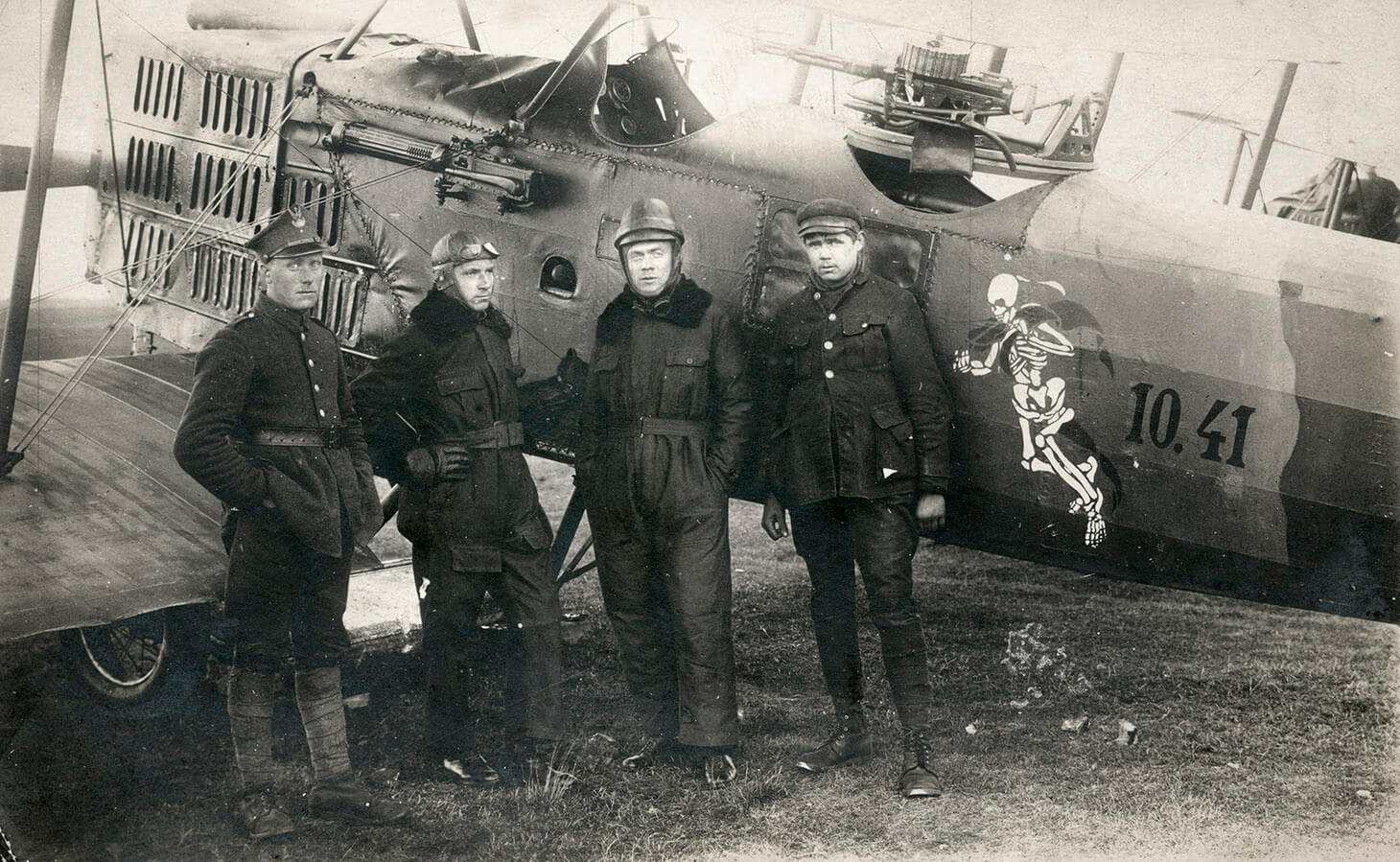 Czarno-białe zdjęcie czterech pilotów na tle samolotu typu Breguet.  Na pierwszym planie, w rzędzie pokazani są w pełnych planach: pierwszy od lewej, żołnierz w mundurze polowym, w lekkim zwrocie w prawo, z prawa ręką w kieszeni spodni. Drugi od lewej stoi w podobnym ustawieniu pilot w kombinezonie lotniczym i pilotce na głowie, podpiera się prawą ręką pod bok. Trzeci od lewej, ustawiony na wprost obiektywu pozuje sierżant Toryślak, w kombinezonie lotniczym, kasku na głowie i z rękami w kieszeniach spodni kombinezonu. Czwarty od lewej, porucznik Ratomski, w kurtce wojskowej, bryczesach i czapce na głowie, z opuszczonymi swobodnie rękami. Samolot będący tłem dla pozujących, skierowany jest w lewą stronę kadru i usytuowany jest równolegle do dolnej, dłuższej krawędzi zdjęcia. Prawa i lewa krawędź zdjęcia przycina nieco ogon samolotu i jego przód, tak że widoczny jest jedynie częściowo dolny płat skrzydeł, kratownica przysłaniająca silnik, kokpit, centralnie działo i fragment ogona. Na boku samolotu, przed ogonem namalowany jest w jasnym kolorze kościotrup i liczby: dziesięć, kropka czterdzieści jeden.