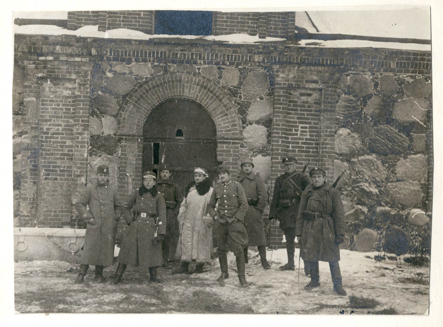 Czarno-biała fotografia przedstawiająca grupę ośmiu żołnierzy pozujących na tle ceglano-kamiennej elewacji magazynu. Za ich plecami masywne, metalowe drzwi zwieńczone  łukiem ceglanym. Mężczyźni w płaszczach mundurowych stoją swobodnie w rzędzie. Dwie centralne postaci wyróżniają się ubraniem. Jeden z żołnierzy bez płaszcza, tylko w mundurze polowym, w bryczesach stoi z rękami w kieszeni. Mężczyzna za nim ubrany jest w ciężki,  jasny kożuch z obszernym futrzanym kołnierzem. Trzej żołnierze po prawej stronie mają przewieszone przez plecy karabiny.