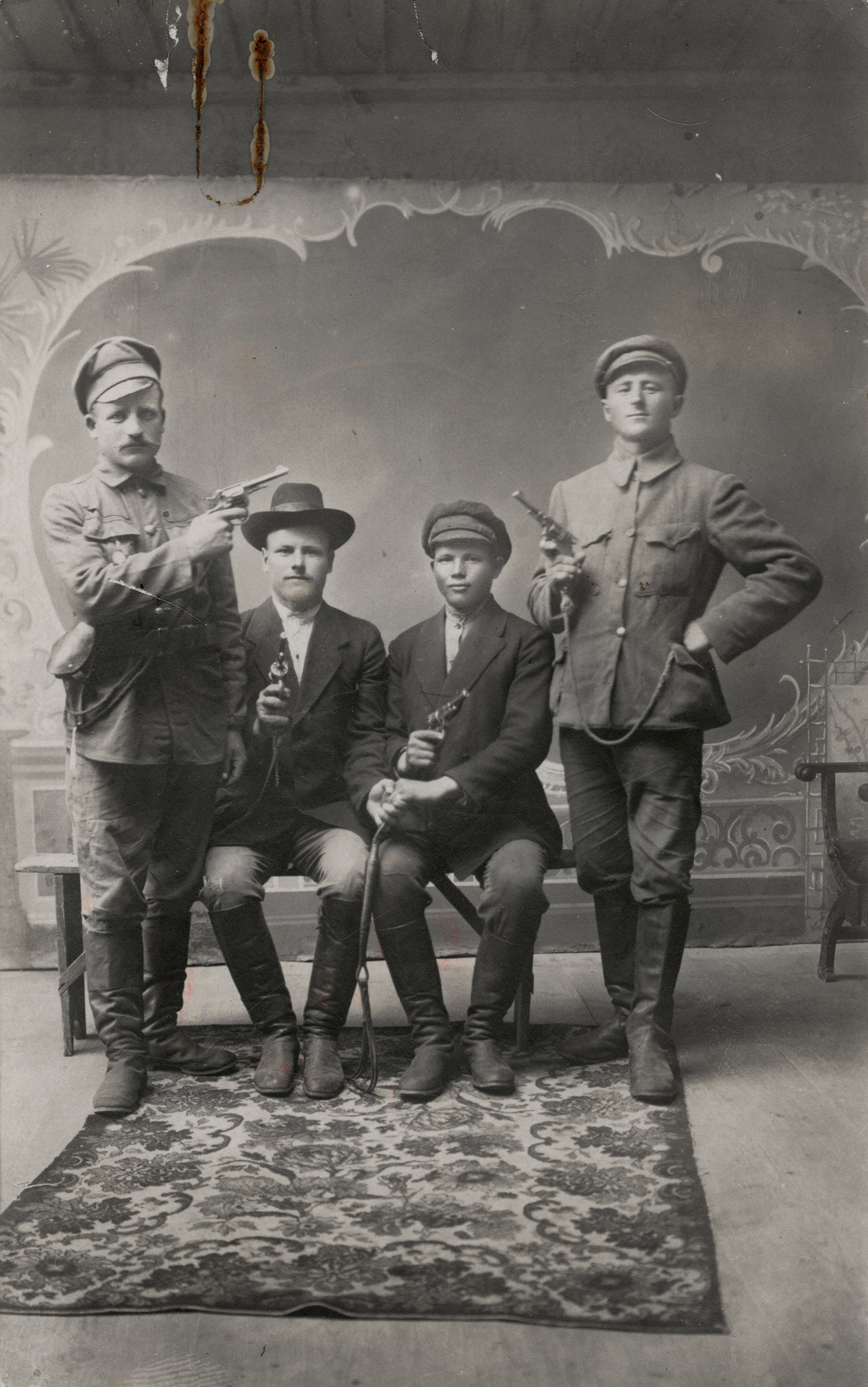 Czarno-biała fotografia studyjna czterech mężczyzn w różnym wieku pozujących z pistoletami. Dwóch z nich, ubranych po cywilnemu, siedzi po środku na prostej, drewnianej ławie, ustawionej na kwiecistym dywanie. Ich lewe dłonie zaciśnięte są razem na elemencie uprzęży końskiej. Po obu ich stronach pozuje, w pozycjach stojących, dwóch mężczyzn w mundurach. Twarze wszystkich zwrócone są w stronę widza. Broń, z której celują wymierzona jest w przypadkowych kierunkach. W tle namalowana na ścianie rama w postaci krętych, fantazyjnych linii. 