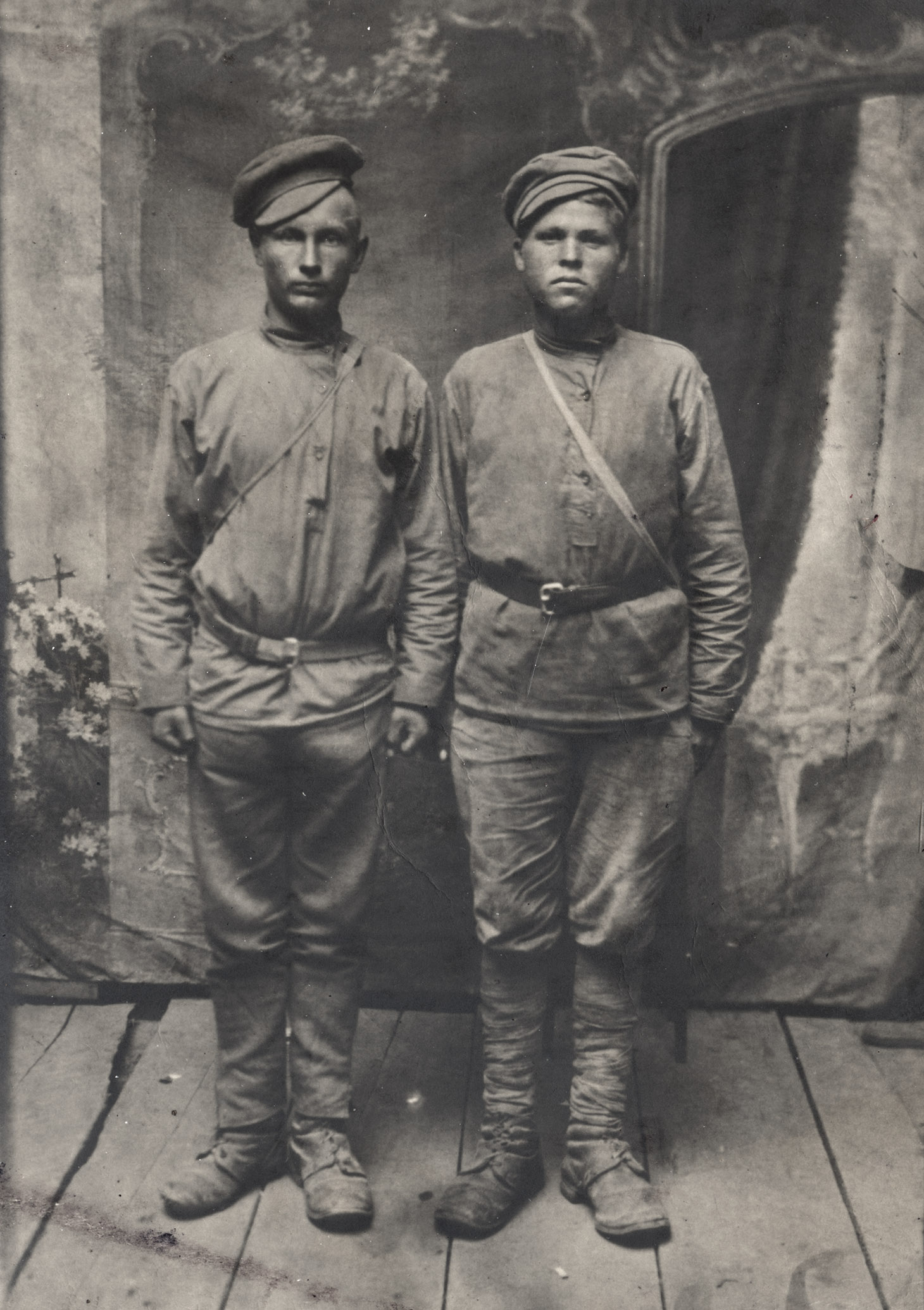 Czarno-biała fotografia studyjna przedstawiająca dwóch mężczyzn młodych mężczyzn w pozycjach "na baczność". Ubrani są w tradycyjne rosyjskie, proste, przepasane koszule z kołnierzem-stójką i długimi rękawami.Mężczyźni stoją wyprostowani w butach z onucami i w czapkach na bakier. Stoją na podłodze z dużych, topornych desek. Tło dla pozujących stanowi dekoracyjna tkanina z elementami florystycznymi i fragment stojącego lustra.