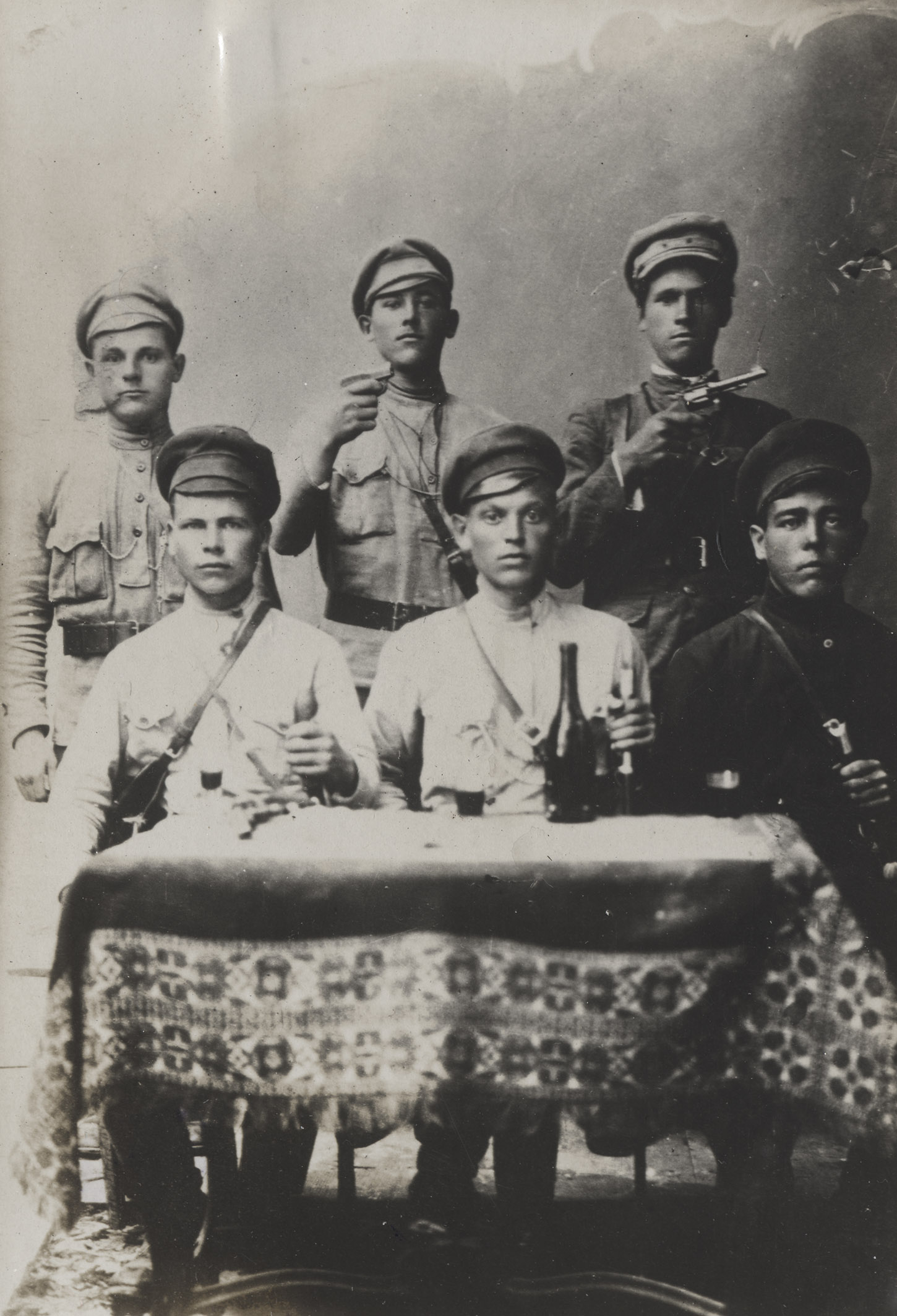 Czarno-biała fotografia sześciu młodych mężczyzn pozujących przy stole we wnętrzu studia. Trzech z nich siedzi za stołem, na wprost widza. Ubrani są w tradycyjne rosyjskie, wojskowe koszule z kołnierzem-stójką, przepasane paskiem i kieszeniami na piersiach. Na głowach mają ciemne czapki. Każdy wspiera rękę na rękojeściach szabli. Za ich plecami stoi trzech pozostałych mężczyzn. Dwóch z nich: drugi i trzeci od lewej w zgiętych, prawych rękach trzymają pistolety, które skierowane są nad głowami siedzących w prawą stronę kadru. Stół, który znajduje się na pierwszym planie jest nakryty grubym, wzorzystym materiałem, a na nim stoi ciemna butelka z długą szyjką oraz trzy szklanki napełnione ciemnym płynem, prawdopodobnie winem.

