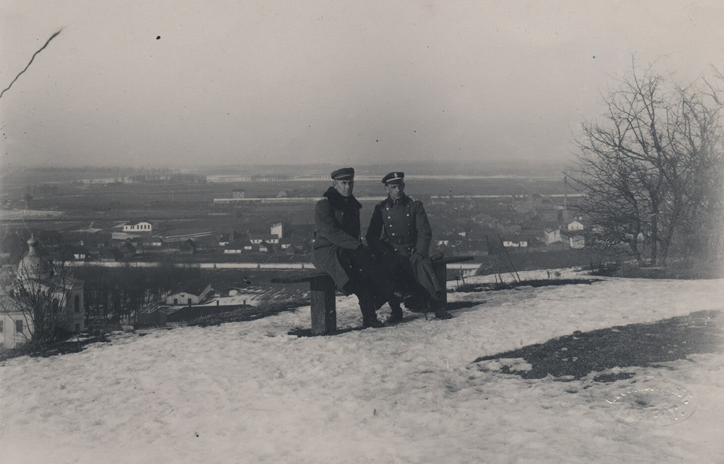 Czarno-biała fotografia, w której  centrum Generał Edward Rydz-Śmigły w towarzystwie oficera siedzi na prostej, drewnianej ławce. Mężczyźni ubrani są w wojskowe, zimowe płaszcze. Przed nimi na trawie zalega topniejący śnieg. Z prawej strony kadru widać konary bezlistnych krzewów. Za plecami siedzących, poniżej punktu widokowego i w dali rozpościera się miasto.