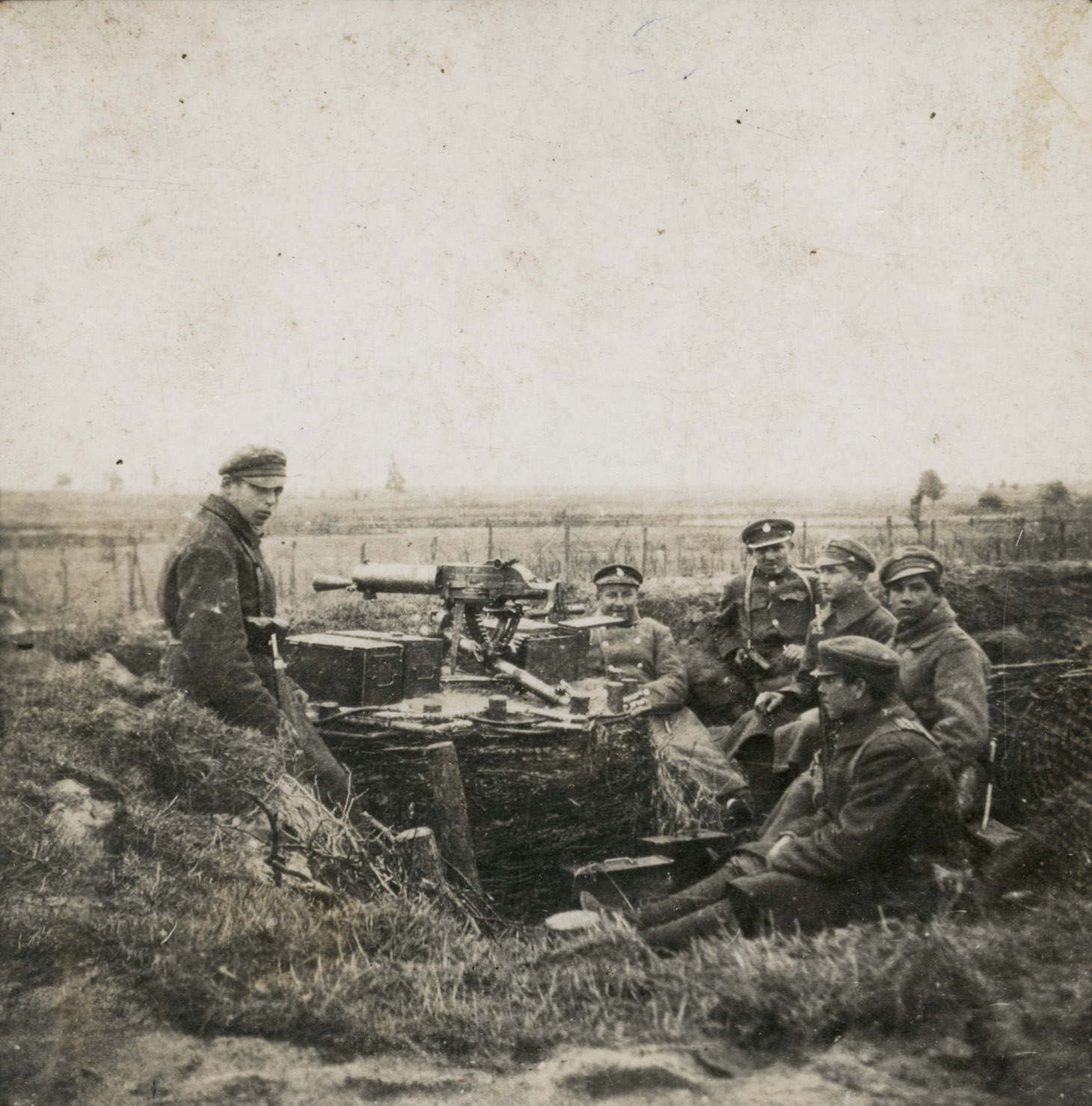 Czarno-biała fotografia przedstawiająca sześciu, pozującymi żołnierzy w okopie, przy stanowisku z karabinem. W dali lekko zaznaczony horyzont pól.  W centrum planu znajduje się karabin skierowany w stronę horyzontu i skrzynie z amunicją. Żołnierze siedzą w swobodnych pozach i opierając się plecami o wał okopu.