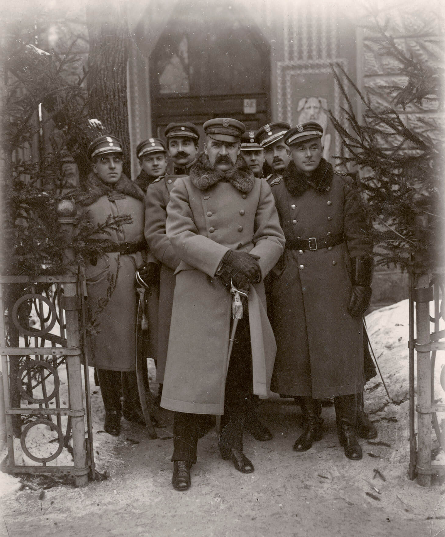 Czarno-biała fotografia przedstawiająca żołnierzy stojących  w swobodnych pozach, w otwartej furtce metalowego ogrodzenia, na tle budynku. Po środku, wysunięty do przodu, stoi Naczelnik Państwa Józef Piłsudski w dwurzędowym płaszczu wojskowym z futrzanym kołnierzem, w czapce "maciejówce", z dłońmi wspartymi o rękojeść szabli. Obok, po prawej stronie, w ciemniejszym, dwurzędowym płaszczu wojskowym z futrzanym kołnierzem spiętym skórzanym paskiem stoi generał Edward Rydz-Śmigły. Pozostałych pięciu żołnierzy na drugim planie ubrani są w płaszcze wojskowe i czapki "maciejówki". Po obu stronach kadru widnieją fragmenty niskiego  ozdobnego, metalowego ogrodzenia i gałęzie świerków.