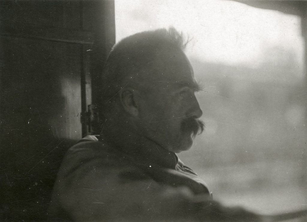 Czarno-biała fotografia przedstawiająca portret Marszałka Józefa Piłsudskiego w pełnym profilu we wnętrzu pociągu, na lekko rozmytym tle pejzażu za oknem.