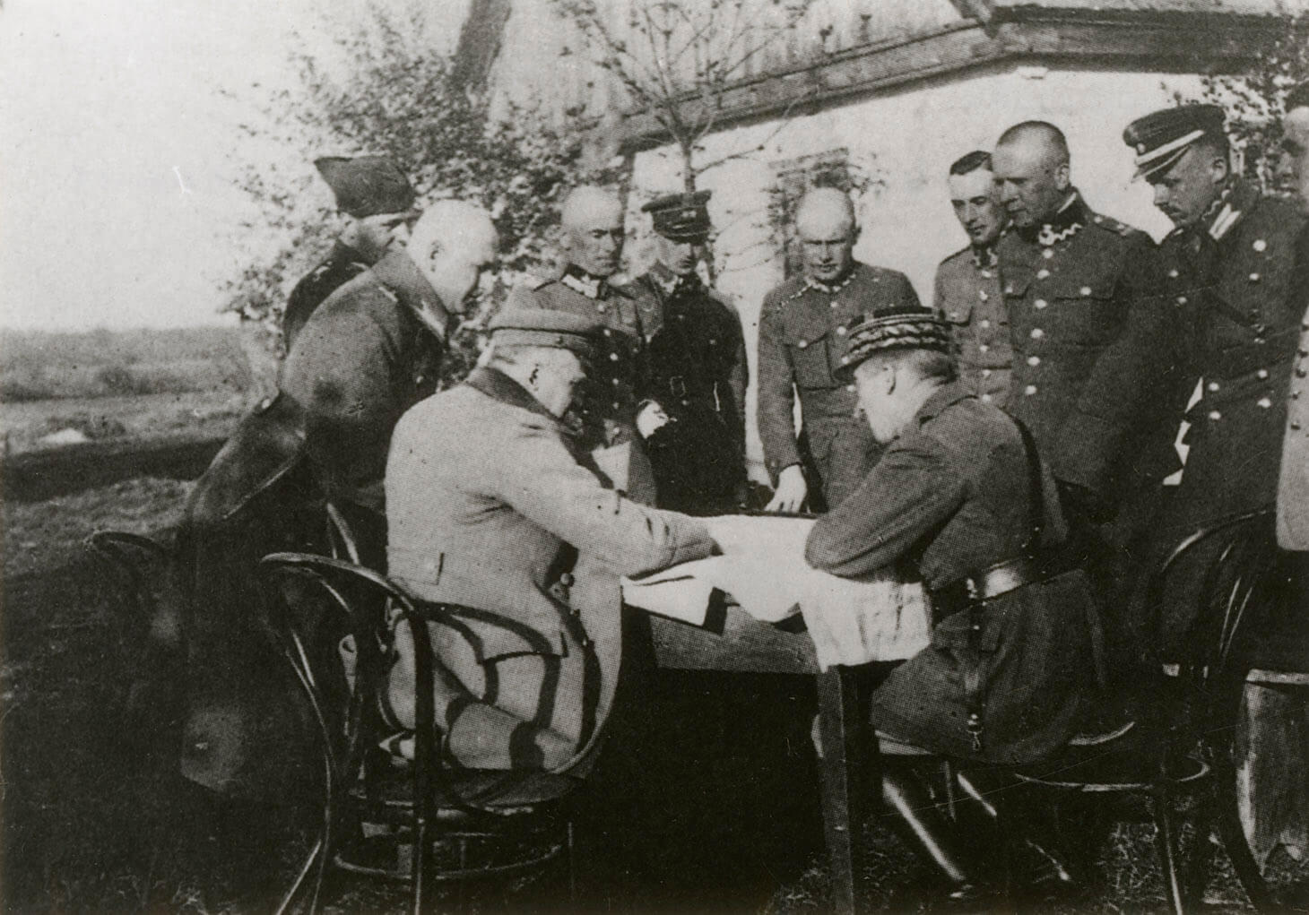 Czarno-biała fotografia przedstawiająca naradę dziewięciu oficerów wokół stołu znajdującego przed drewnianym wiejskim domem. Za nim, po lewej stronie widoczny horyzont pól. Krąg oficerów zamykają dwie główne postaci siedzące na pierwszym planie, profilami do widza. Pierwszy z lewej to Naczelny Wódz Józef Piłsudski, drugi od lewej to generał Paul Henrys. Obydwaj mężczyźni siedzą na krzesłach w stylu Thoneta, pochyleni są nad mapą rozłożoną na stole. Marszałek ubrany jest w rozpięty, jasny wojskowy płaszcz, na głowie ma czapkę maciejówkę. Generał Paul Henrys, ma na sobie mundur przepasany pasem oficerskim z koalicyjką - paskiem założonym na skos w poprzek piersi i przytroczonym do pasa głównego. Po lewej stronie Marszałka Piłsudskiego, stoi w wojskowym płaszczu, bez nakrycia głowy, Generał Edward Śmigły-Rydz. 
