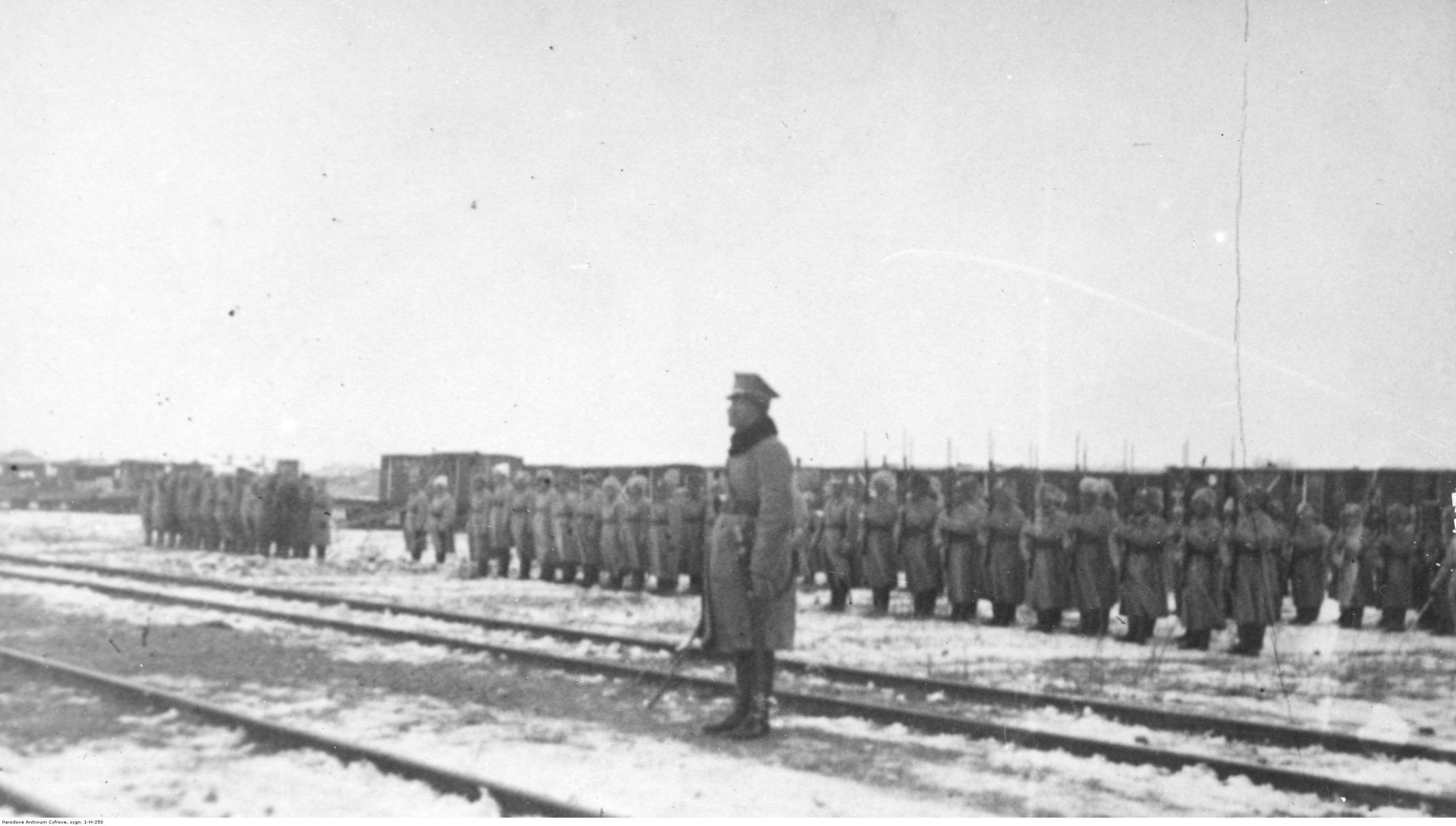 Czarno-biała fotografia przedstawiająca żołnierzy Dywizji Syberyjskiej ustawionej w szeregach, pośród torów kolejowych. Wszyscy w długich, zimowych płaszczach i futrzanych czapach.  W centrum kadru znajduje się żołnierz w zimowym, płaszczu wojskowym. Stoi lewym bokiem do obiektywu, w postawie zasadniczej z szablą opuszczoną swobodnie w prawej ręce. Spogląda w swoją prawą stronę. 